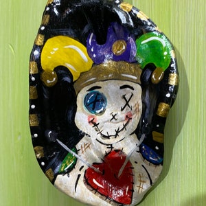 May include: Hand-painted, decorative oyster shell featuring a voodoo doll design. The doll has a stitched face, a red heart, and is adorned with a jester's hat in yellow, purple, and green. The shell is edged with gold and black accents.