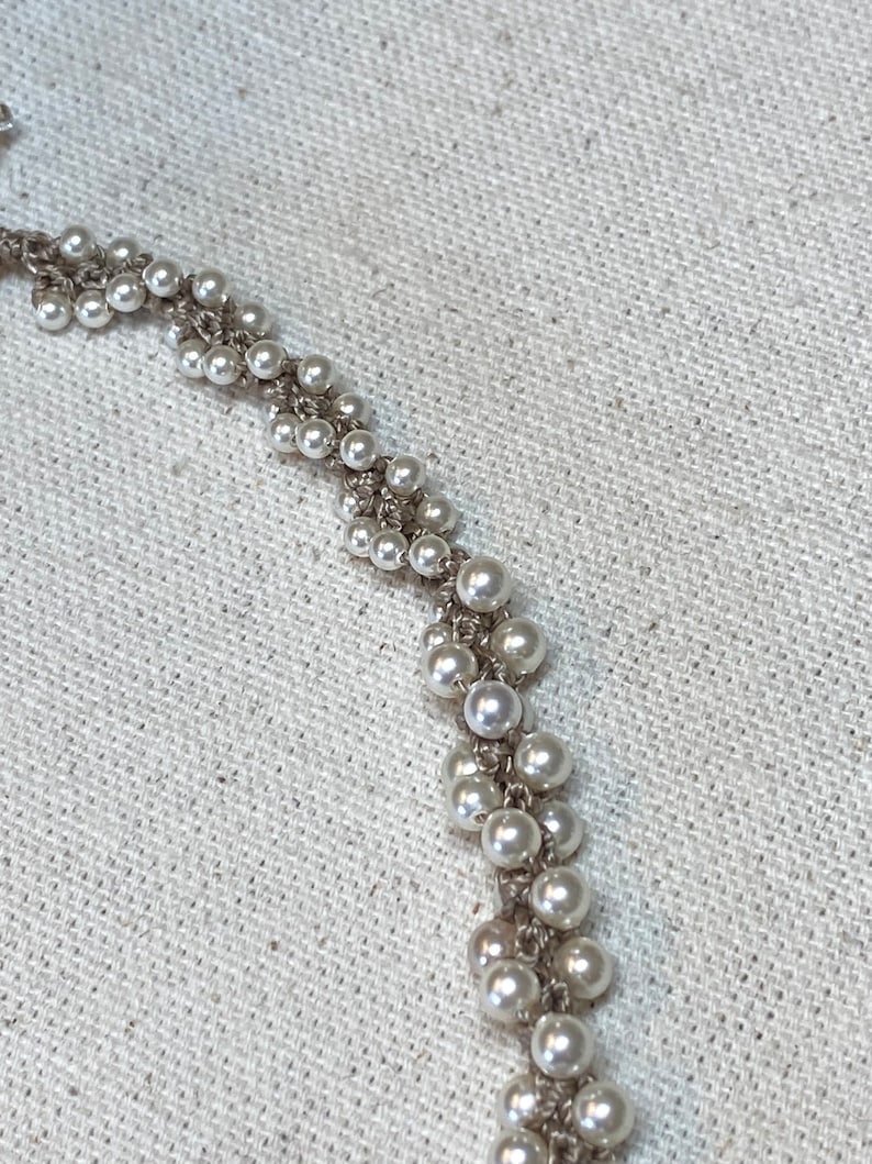 May include: A close-up of a white pearl necklace with a delicate chain design. The necklace is made of small, round pearls that are strung together in a repeating pattern.
