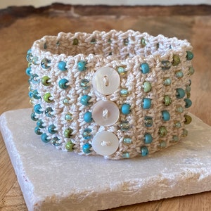 K&ouml;nnte beinhalten: Ein geh&auml;keltes Armband mit beigefarbener Basis und blauen und gr&uuml;nen Perlen. Das Armband hat drei wei&szlig;e Kn&ouml;pfe.