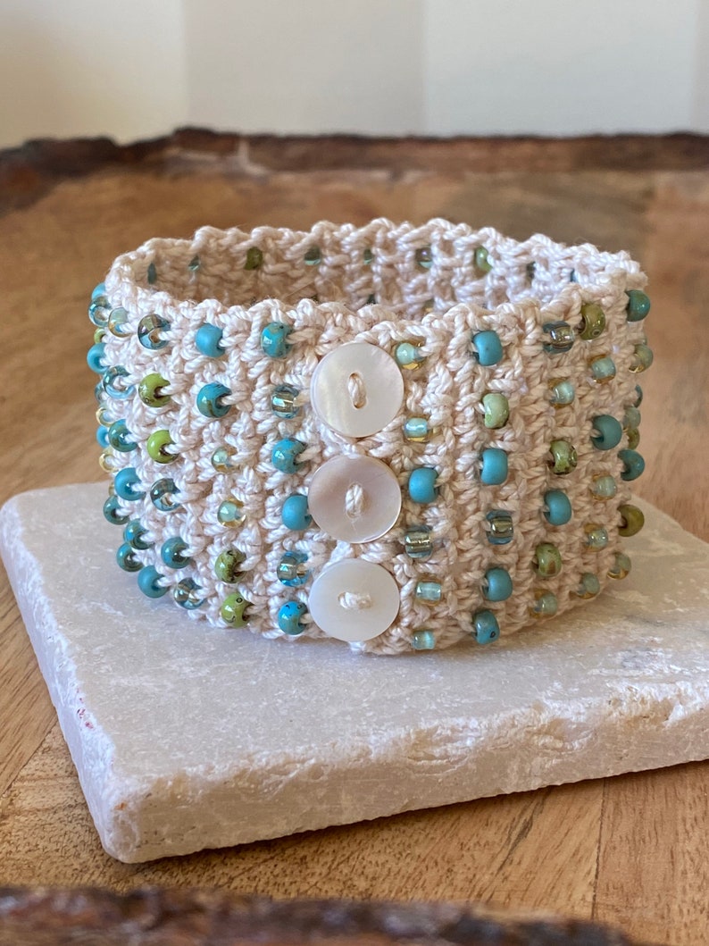 K&ouml;nnte beinhalten: Ein geh&auml;keltes Armband mit beigefarbener Basis und blauen und gr&uuml;nen Perlen. Das Armband hat drei wei&szlig;e Kn&ouml;pfe.