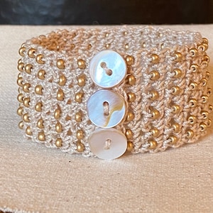 K&ouml;nnte beinhalten: Ein beigefarbenes geh&auml;keltes Armband mit goldenen Perlen und drei wei&szlig;en Muschelknäpfen.