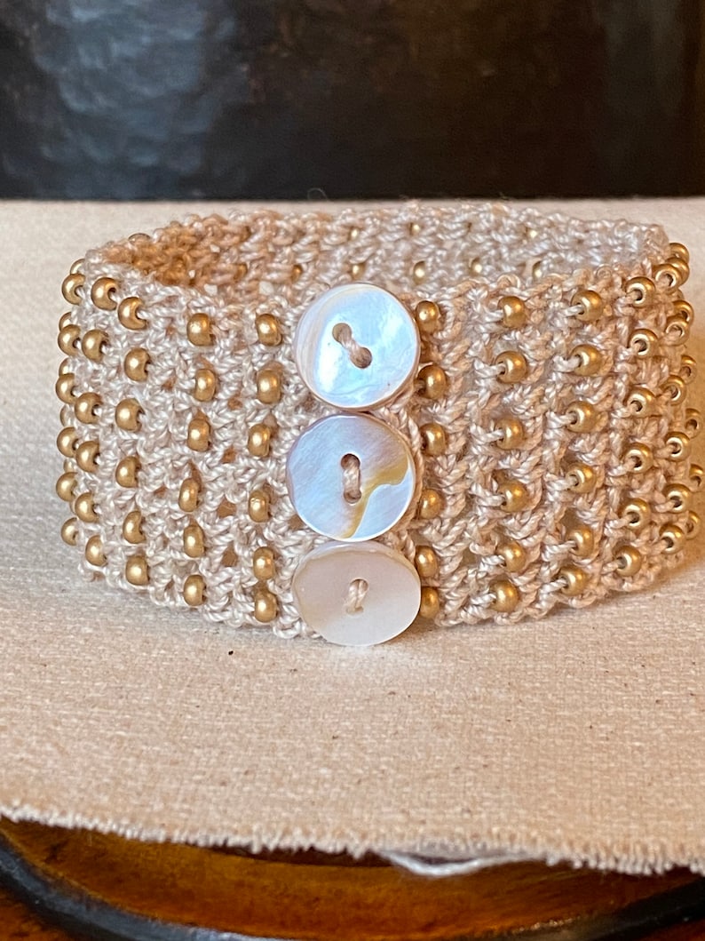 K&ouml;nnte beinhalten: Ein beigefarbenes geh&auml;keltes Armband mit goldenen Perlen und drei wei&szlig;en Muschelknäpfen.