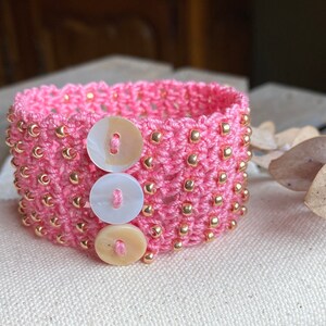 K&ouml;nnte beinhalten: Rosa geh&auml;keltes Armband mit goldenen Perlen und drei wei&szlig;en und cremefarbenen Kn&ouml;pfen.
