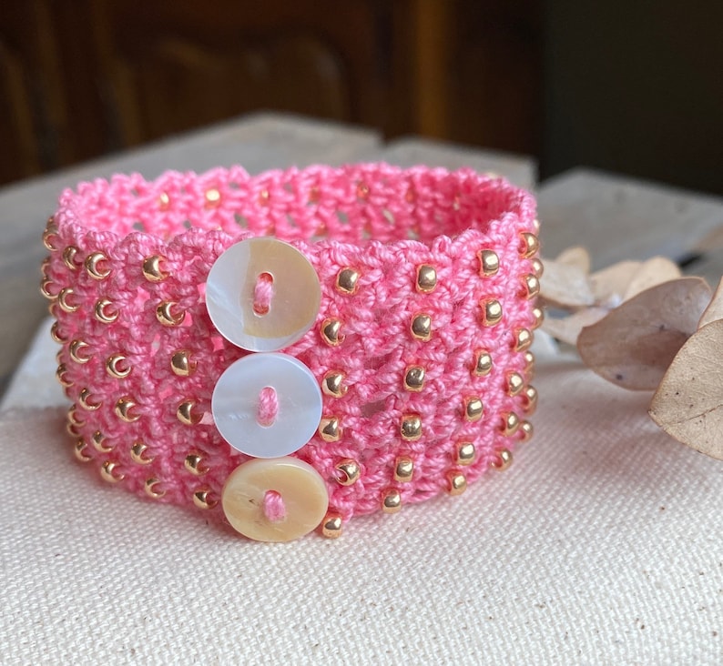 K&ouml;nnte beinhalten: Rosa geh&auml;keltes Armband mit goldenen Perlen und drei wei&szlig;en und cremefarbenen Kn&ouml;pfen.