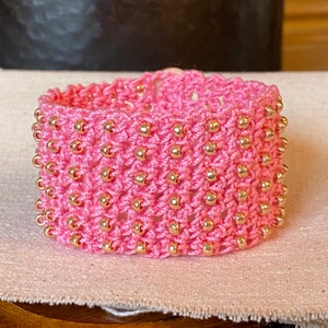 K&ouml;nnte beinhalten: Ein pinkfarbenes geh&auml;keltes Armband mit goldenen Perlen. Das Armband ist mit einem gewebten Muster gearbeitet und hat einen zarten, femininen Look.