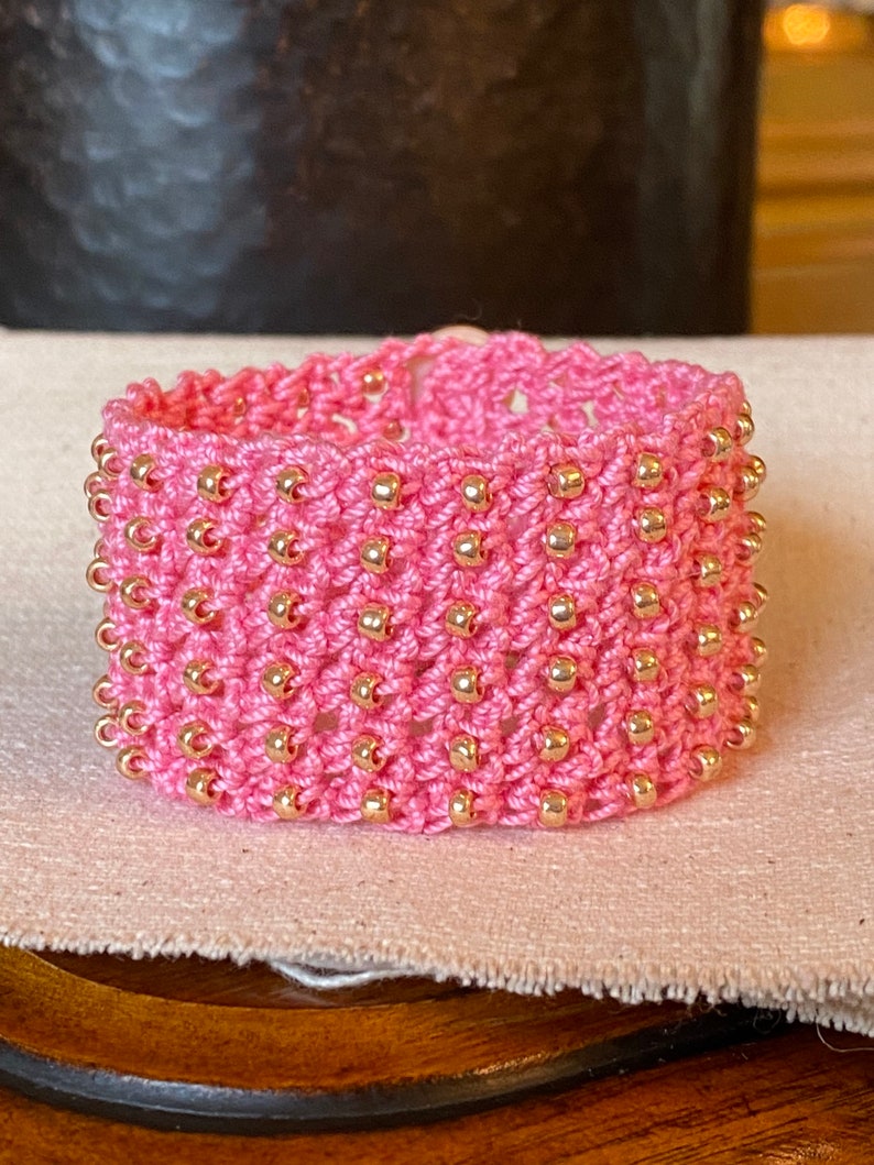 K&ouml;nnte beinhalten: Ein pinkfarbenes geh&auml;keltes Armband mit goldenen Perlen. Das Armband ist mit einem gewebten Muster gearbeitet und hat einen zarten, femininen Look.