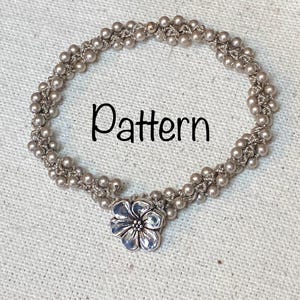 Puede incluir: Una pulsera plateada con un cierre en forma de flor. La pulsera está hecha de pequeñas cuentas redondas y presenta la palabra "Pattern" en escritura negra. Se muestra sobre una superficie texturizada de color blanquecino.
