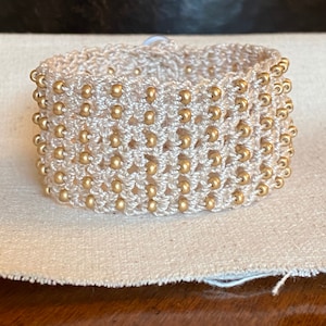 K&ouml;nnte beinhalten: Ein wei&szlig;es geh&auml;keltes Armband mit goldenen Perlen. Das Armband ist mit einer einfachen Masche gearbeitet und hat einen zarten, femininen Look.