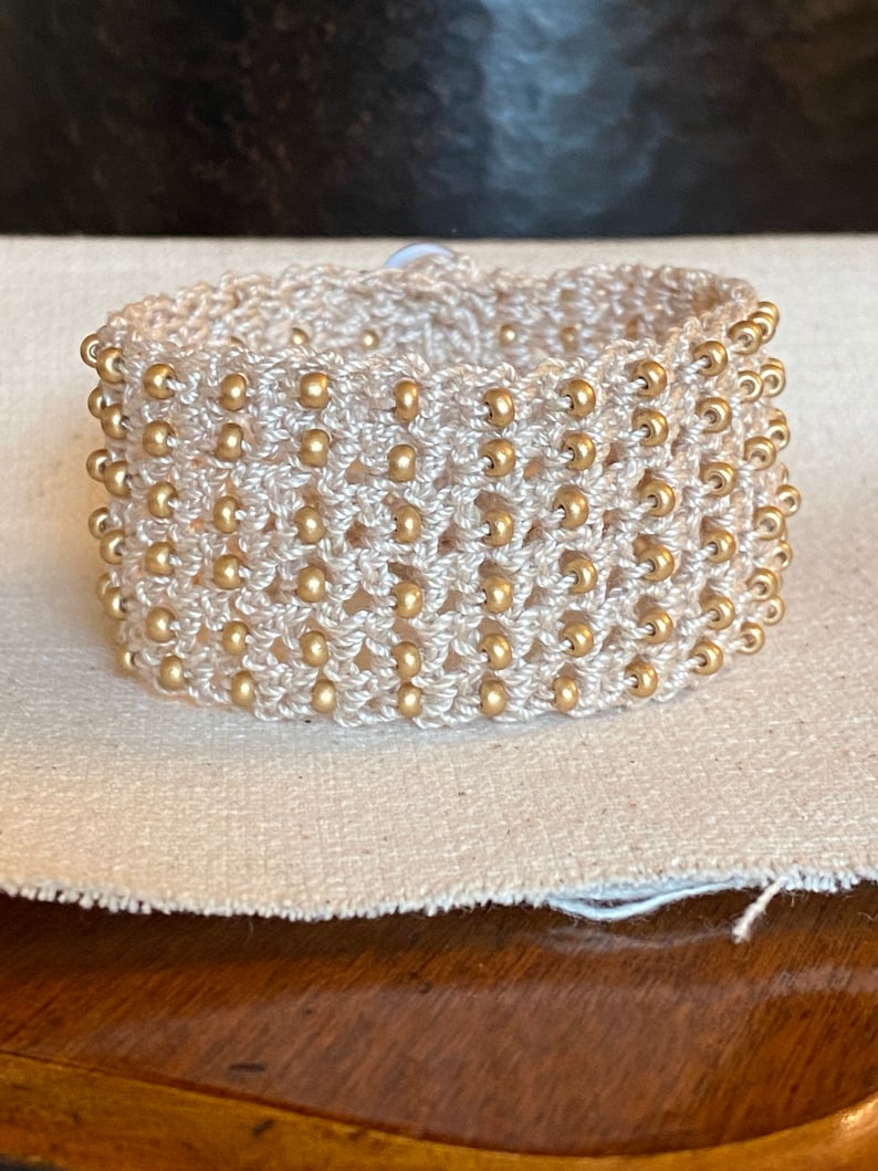 K&ouml;nnte beinhalten: Ein wei&szlig;es geh&auml;keltes Armband mit goldenen Perlen. Das Armband ist mit einer einfachen Masche gearbeitet und hat einen zarten, femininen Look.