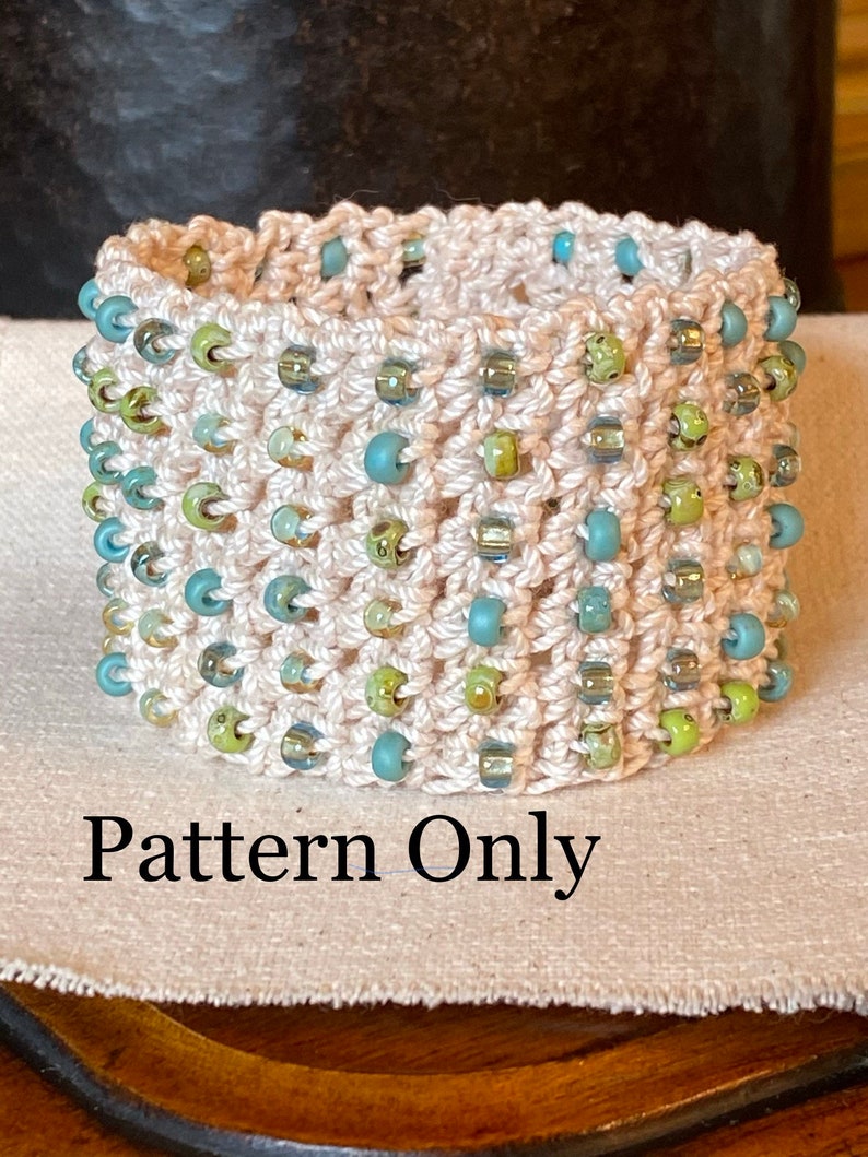 K&ouml;nnte beinhalten: Ein geh&auml;keltes Armband mit einer wei&szlig;en Basis und blauen, gr&uuml;nen und silbernen Perlen. Das Armband liegt auf einer beigefarbenen Stoffoberfl&auml;che. Nur das Muster.