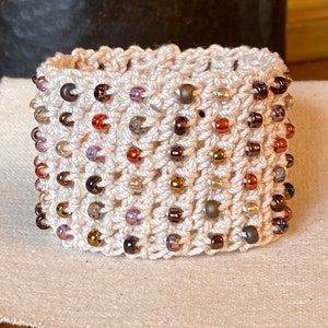 K&ouml;nnte beinhalten: Ein geh&auml;keltes Armband mit cremefarbener Basis und einer Vielzahl von braunen, goldenen und violetten Perlen.