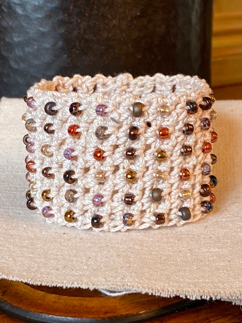 K&ouml;nnte beinhalten: Ein geh&auml;keltes Armband mit cremefarbener Basis und einer Vielzahl von braunen, goldenen und violetten Perlen.