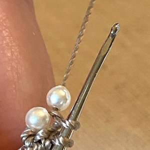 May include: A close-up of a crochet hook with two white beads on the cord. The hook is made of metal and has a silver finish.