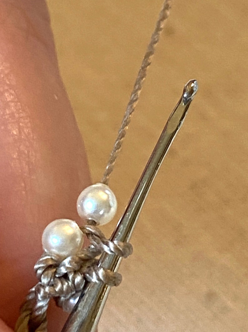 May include: A close-up of a crochet hook with two white beads on the cord. The hook is made of metal and has a silver finish.