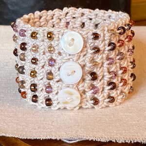 K&ouml;nnte beinhalten: Ein geh&auml;keltes Armband mit braunen und violetten Perlen und drei wei&szlig;en Kn&ouml;pfen. Das Armband liegt auf einer beigefarbenen Stoffoberfl&auml;che.
