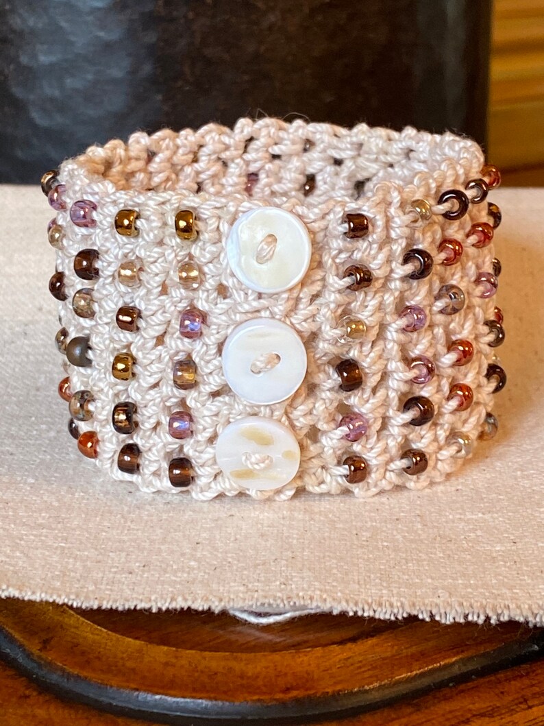 K&ouml;nnte beinhalten: Ein geh&auml;keltes Armband mit braunen und violetten Perlen und drei wei&szlig;en Kn&ouml;pfen. Das Armband liegt auf einer beigefarbenen Stoffoberfl&auml;che.