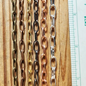 May include: Seven different colored metal chains with oval links. The chains are hanging from a wooden surface with a ruler in the background. The ruler is marked in inches.