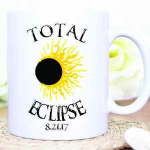 May include: White ceramic mug featuring a solar eclipse design. The design includes a black circle representing the moon, surrounded by a yellow sun with the words "TOTAL ECLIPSE" and the date "8.21.17" in black text.