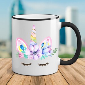 May include: White ceramic mug with a black rim and handle. The mug features a watercolor illustration of a unicorn's face with closed eyes, a horn, and floral accents in pink, purple, blue, and green.