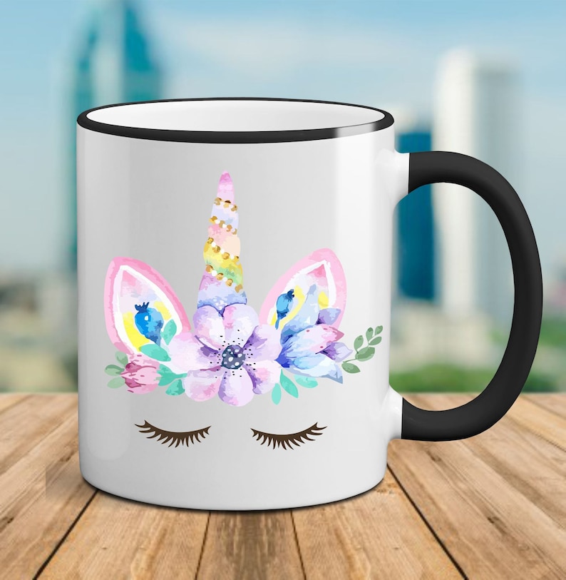 May include: White ceramic mug with a black rim and handle. The mug features a watercolor illustration of a unicorn's face with closed eyes, a horn, and floral accents in pink, purple, blue, and green.
