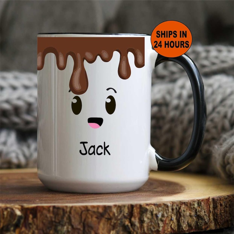 K&ouml;nnte beinhalten: Wei&szlig;e Keramik-Tasse mit schwarzem Henkel und Rand, mit einem Cartoon-Marshmallow-Gesicht mit brauner Schokolade, die von oben tropft. Der Name "Jack" ist darunter aufgedruckt.