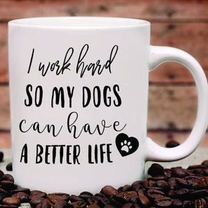 May include: White ceramic coffee mug with black text that reads "I work hard so my dogs can have a better life" with a black paw print inside a heart.