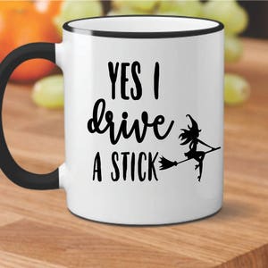 Op de afbeelding: Een witte keramische mok met een zwarte rand en handvat. De mok heeft een zwarte afbeelding van een heks die op een bezem vliegt met de tekst "YES I drive a STICK".
