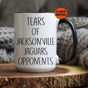 May include: A white ceramic mug with a black handle and rim. The front of the mug features black text that reads: TEARS OF JACKSONVILLE JAGUARS OPPONENTS. A small orange circular badge in the upper right corner contains the text 24 HOUR SHIPPING.