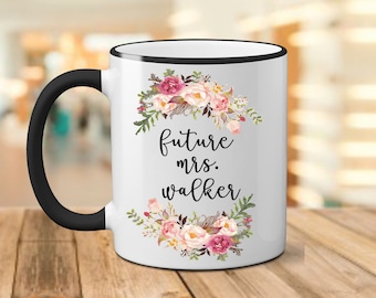 May include: White ceramic mug with a black rim and handle. The mug has a floral design with pink, white, and green flowers. The text "future mrs. walker" is printed in black on the mug.