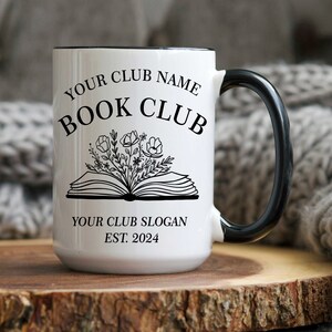 May include: White ceramic mug with a black rim and handle. The mug has a black and white design of an open book with flowers and the text "YOUR CLUB NAME BOOK CLUB YOUR CLUB SLOGAN EST. 2024".
