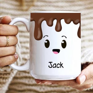 K&ouml;nnte beinhalten: Wei&szlig;e Keramik-Tasse mit einem Cartoon-Marshmallow-Design. Die Oberseite der Tasse zeigt ein braunes Schokoladen-Tropf-Design mit einem l&auml;chelnden Marshmallow-Gesicht darunter. Der Name "Jack" ist in schwarzer Schrift auf die Tasse gedruckt.