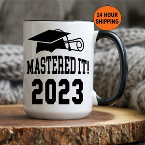 May include: White ceramic mug with a black handle and rim. The mug features a graduation cap and diploma graphic, along with the text "MASTERED IT! 2023" in bold black letters. A graduation-themed gift.