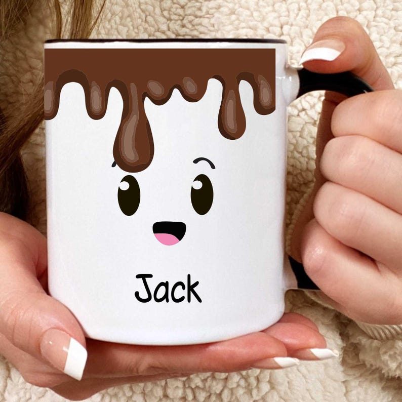 K&ouml;nnte beinhalten: Wei&szlig;e Keramik-Tasse mit schwarzem Henkel und Rand, mit einem Cartoon-Gesicht mit Schokolade, die von oben tropft. Die Tasse hat ein l&auml;chelndes Gesicht mit dem Namen "Jack" darunter. Die Tasse wird von einer Person gehalten.