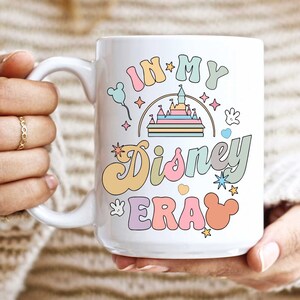 Op de afbeelding: Witte keramische mok met een kleurrijk grafisch ontwerp met de tekst "In My Disney Era". Het ontwerp bevat een cartoonkasteel, Mickey Mouse-oren en sterren.