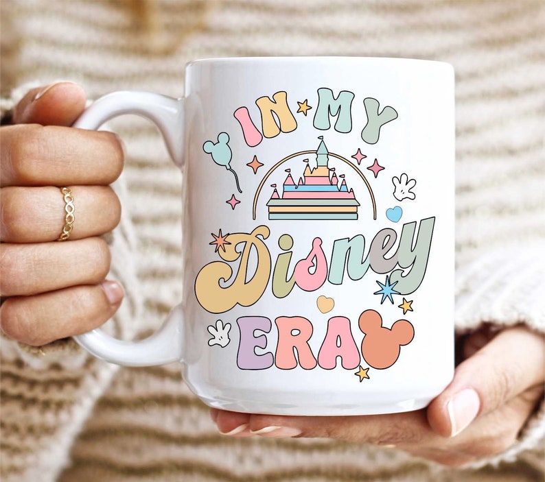 Op de afbeelding: Witte keramische mok met een kleurrijk grafisch ontwerp met de tekst "In My Disney Era". Het ontwerp bevat een cartoonkasteel, Mickey Mouse-oren en sterren.