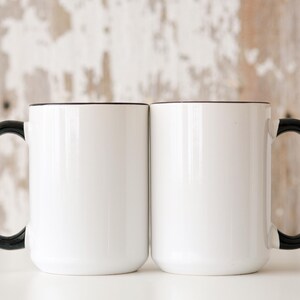 May include: Two white ceramic mugs with black handles and a black rim. The mugs are empty and sitting on a white surface.