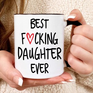 May include: White ceramic coffee mug with a black handle. The mug has black text that reads "BEST F♡CKING DAUGHTER EVER".