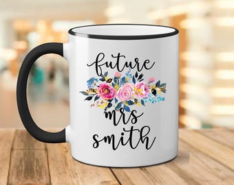 May include: White ceramic mug with a black rim and handle. The mug has a floral design and the text "future mrs smith" in black.