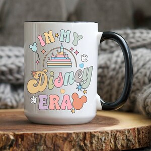 Op de afbeelding: Witte keramische mok met een zwart handvat. De mok heeft een kleurrijke afbeelding met de tekst "In My Disney Era" met een cartoonkasteel, sterren en Mickey Mouse-oren.