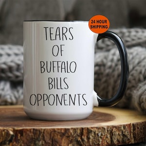 May include: White ceramic coffee mug with a black handle and black rim. The mug has black text that reads "Tears of Buffalo Bills Opponents".