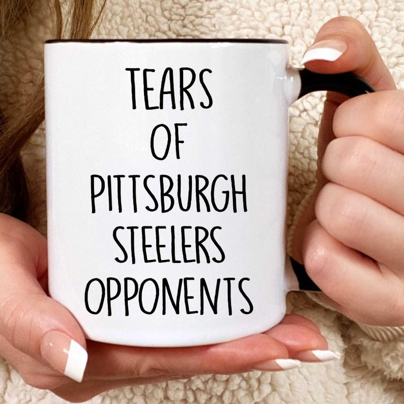 K&ouml;nnte beinhalten: Eine wei&szlig;e Keramik-Kaffeetasse mit schwarzem Rand und Henkel. Die Tasse hat einen schwarzen Schriftzug mit der Aufschrift "Tears of Pittsburgh Steelers Opponents".