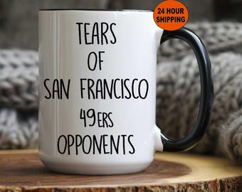 Op de afbeelding: Een witte keramische koffiemok met een zwarte handgreep en een zwarte rand. De mok heeft zwarte tekst die "Tears of San Francisco 49ers Opponents" luidt.
