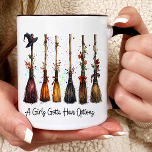 May include: White ceramic mug with a black handle and rim, featuring a design of six decorative witch brooms in various colors. The brooms are adorned with leaves and bows, and the text "A Girls Gotta Have Options" is printed on the mug.