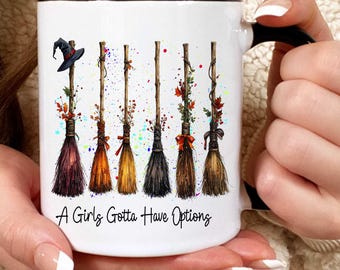 May include: White ceramic mug with a black handle and rim, featuring a design of six decorative witch brooms in various colours. The brooms are adorned with leaves and bows, and the text "A Girls Gotta Have Options" is printed on the mug.