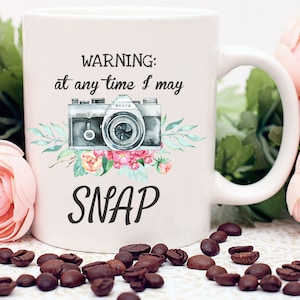 May include: White ceramic mug with a black and white camera illustration and pink flowers. The mug has the text "WARNING: at any time I may SNAP" printed on it.