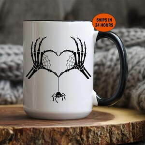 May include: White ceramic mug with a black handle and rim. The mug features a black skeleton hand design forming a heart shape with a spider and web detail. The text "SHIPS IN 24 HOURS" is on the top right.