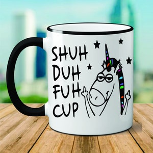 May include: White ceramic coffee mug with a black rim and handle. The mug features a black and white illustration of a unicorn with a rainbow mane giving the middle finger. The text "SHUH DUH FUH CUP" is printed in black on the mug.