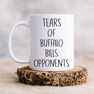 May include: White ceramic coffee mug with black text that reads "Tears of Buffalo Bills Opponents". The mug is sitting on a wooden coaster with moss.
