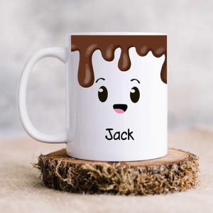 K&ouml;nnte beinhalten: Wei&szlig;e Keramik-Tasse mit einem Cartoon-Marshmallow-Gesicht. Die Tasse hat ein braunes Schokoladen-Tropf-Design oben, mit dem Namen "Jack" in Schwarz darunter gedruckt. Die Tasse steht auf einem Holzuntersetzer.