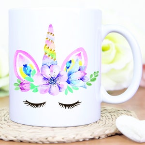May include: White ceramic mug with a watercolor unicorn design. The unicorn has a pink and yellow mane, a gold horn, and closed eyes. The unicorn is surrounded by pink, purple, and blue flowers.
